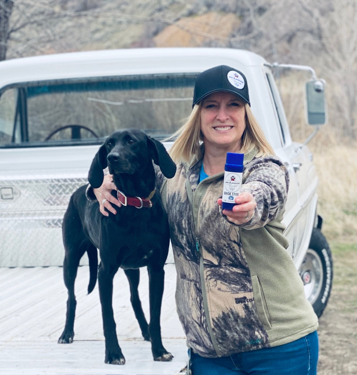 Black dog and a blond female holding a push up tube of Paws R Protected, This product is used for pre-conditioning animal paws and skin, repairing damage of animal paws or skin, and promoting healing of the same. Old white Ford truck.