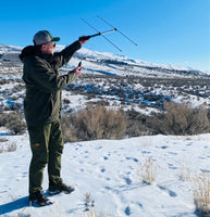 Man standing on a snowy mountain hillside pointing the Beamer Antenna in the direction of where he thinks his dogs might be. The sky is blue and sun is shining. Man is wearing a winter coat and ball cap, holding a GPS antenna.