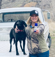 Black dog and a blond female holding a push up tube of Paws R Protected, This product is used for pre-conditioning animal paws and skin, repairing damage of animal paws or skin, and promoting healing of the same. Old white Ford truck.